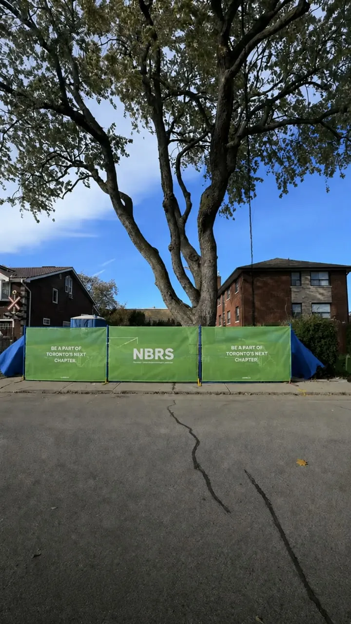 Fenced Toronto infill site with NBRS construction signage and a mature tree